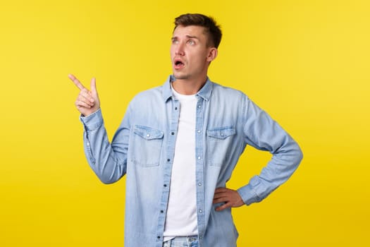 Confused and surprised startled handsome adult man asking question about product, looking and pointing finger upper left corner, consulting shop clerk, standing yellow background.
