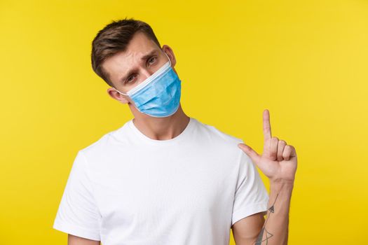 Concept of social distancing, covid-19 and people emotions. Tired gloomy and upset young man in medical mask, looking reluctant at camera as pointing finger up, yellow background.