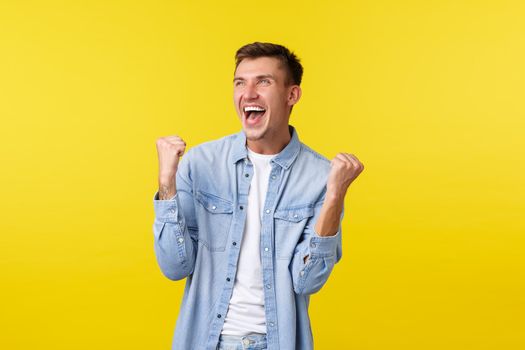 Lifestyle, people emotions and summer leisure concept. Happy handsome blond man raising hands up and shouting yes, feeling rejoice as triumphing, winning bet, gambling over yellow background.