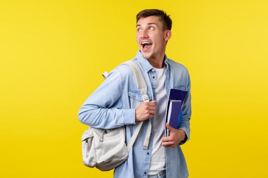 Education, courses and university concept. Excited happy smiling handsome student turn back at upper left corner while heading to classes with backpack and notebooks, yellow background.