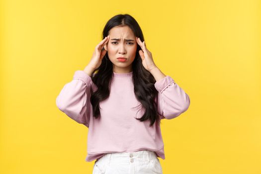People emotions, lifestyle and fashion concept. Distressed and exhausted asian woman close eyes and touching temples, having migraine, feeling headache or dizzy, yellow background.