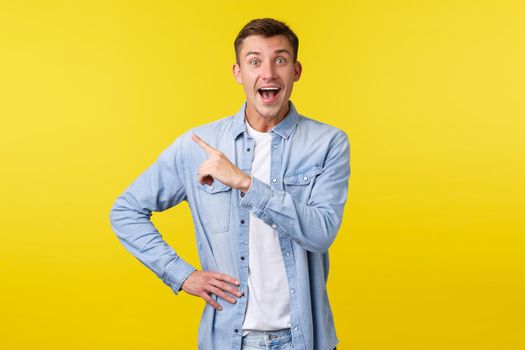 Enthusiastic happy handsome blond man in stylish outfit, pointing finger upper left corner and smiling broadly, found out amazing great news, showing special offer banner, yellow background.