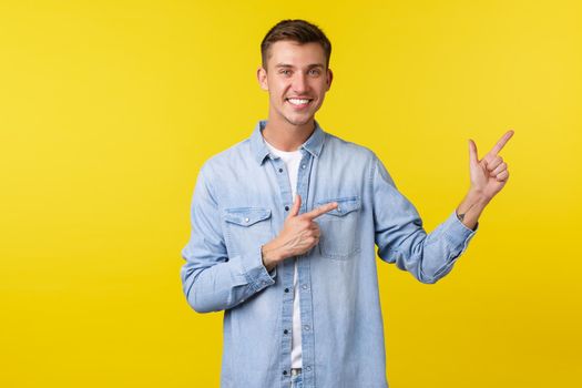 Handsome smiling happy man with white teeth, pointing fingers right, inviting customers check out advertisement, demonstrate new product, special discount offers, standing yellow background.