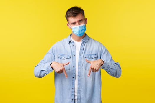 Concept of covid-19 pandemic outbreak, lifestyle during coronavirus social distancing. Skeptical and disappointed man in medical mask tilt head and look bothered while pointing down at banner.