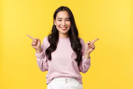 People emotions, lifestyle and fashion concept. Cheerful asian woman smiling and showing both products, pointing fingers sideways at two offers or banners over yellow background.