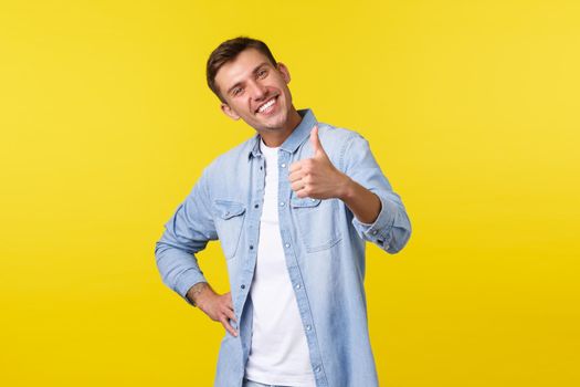 Portrait of satisfied proud young handsome man congratulate person for accomplishing goal, show thumbs-up delighted, smiling pleased. Man recommending awesome offer, stand yellow background.