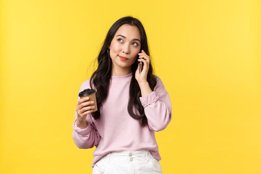 People emotions, lifestyle leisure and beauty concept. Cute stylish asian woman looking up thoughtful as talking on mobile phone and drinking coffee from takeaway cup, yellow background.