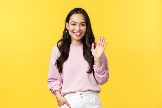 Lifestyle, emotions and advertisement concept. Cute stylish asian girlfriend waving hand to say hi, smiling friendly as greeting someone, make hello or welcome gesture, yellow background.