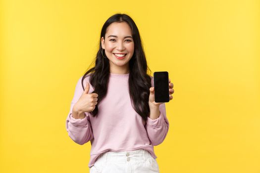 People emotions, lifestyle leisure and beauty concept. Smiling asian woman 20s, showing smartphone display, recommend application or mobile game, showing thumbs-up in approval, yellow background.