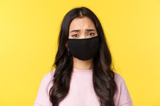 Covid-19, social-distancing lifestyle, prevent virus spread concept. Pity and sad asian gloomy girl frowning, wear face mask, feeling compassion and sadness, standing yellow background.