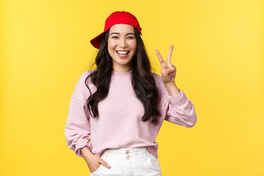 People emotions, lifestyle leisure and beauty concept. Carefree happy, asian hipster girl in summer cap, showing peace sign and smiling, enjoying sunny weekend over yellow background.