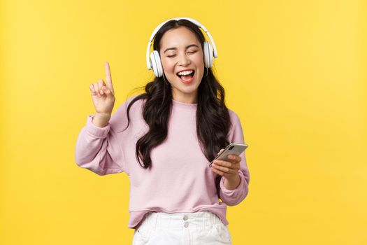 People emotions, lifestyle leisure and beauty concept. Carefree happy asian woman listening music in wireless headphones, holding mobile phone, singing along favorite song, yellow background.