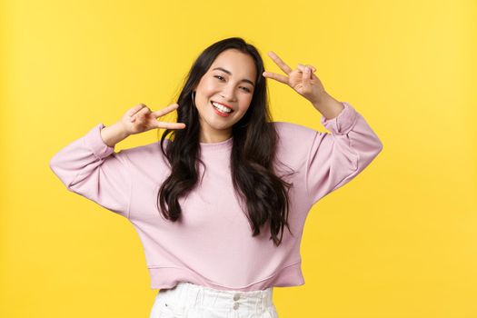 People emotions, lifestyle leisure and beauty concept. Cheerful and optimistic asian girl in stylish clothes, showing peace signs and smiling broadly, enjoying summer, yellow background.