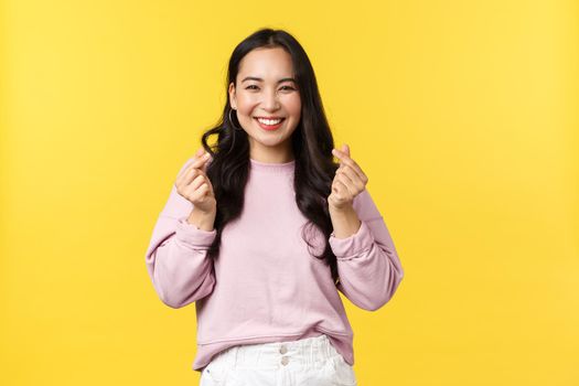 People emotions, lifestyle and fashion concept. Kawaii asian girl with beautiful smile showing korean hearts gesture sending positive and joyful vibes, standing yellow advertisement background.