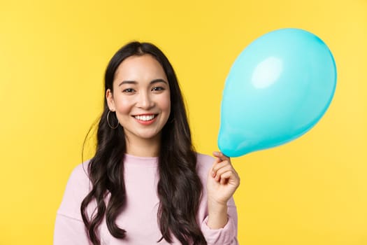 People emotions, lifestyle leisure and beauty concept. Cute smiling asian girl holding blue balloon as congratulating with big event, standing yellow background, through up surprise party.