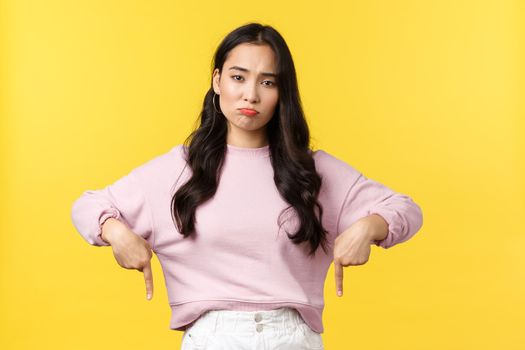 People emotions, lifestyle and fashion concept. Disappointed moody asian girl pointing fingers down and grimacing unsatisfied as showing bad product, complaining, standing yellow background.