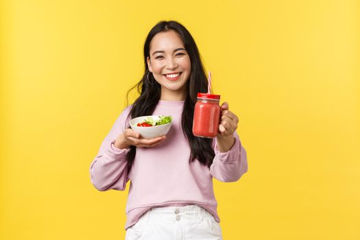 People emotions, healthy lifestyle and food concept. Friendly and cheerful smiling asian girl extend hand and give you smoothie, holding salad, sharing her lunch to eat together, yellow background.