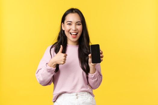 People emotions, lifestyle leisure and beauty concept. Smiling asian woman 20s, showing smartphone display, recommend application or mobile game, showing thumbs-up in approval, yellow background.