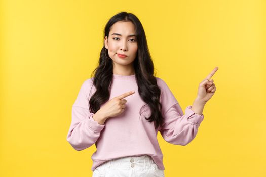 People emotions, lifestyle and fashion concept. Skeptical unimpressed asian woman smirk and looking unbothered as pointing fingers right, standing yellow background. Copy space