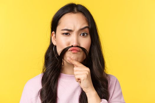 People emotions, lifestyle leisure and beauty concept. Thoughtful funny girl making fake moustache from hair and looking judgemental or suspicious camera, thinking, yellow background.