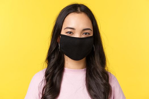 Covid-19, social-distancing lifestyle, prevent virus spread concept. Close-up of young smiling asian girl in face mask standing over yellow background, summer in city, yellow background.