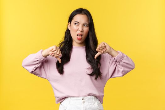 People emotions, lifestyle and fashion concept. Upset and disappointed asian girl pouting and showing disapproval, make thumbs-down in dislike, dont like idea, yellow background.