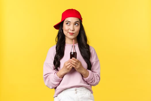 People emotions, drinks and summer leisure concept. Creative stylish asian girl in red cap, smiling intrigued as looking top left corner, drinking soda, enjoying cold beverage, yellow background.
