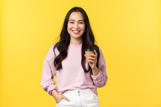 People emotions, lifestyle leisure and beauty concept. Young happy asian female student grab coffee before lecture starts in university, holding cup and laughing at camera carefree, yellow background.