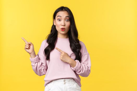People emotions, lifestyle and fashion concept. Surprised and excited stylish asian woman pointing upper left corner at promo banner over yellow background, girl showing cool advertisement.