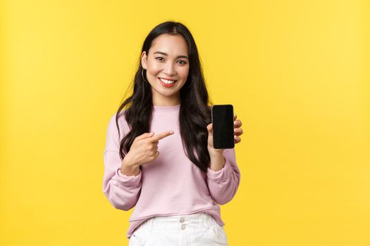 People emotions, lifestyle leisure and beauty concept. Smiling asian woman 20s, showing smartphone display, recommend application or mobile game, pointing finger at screen, yellow background.