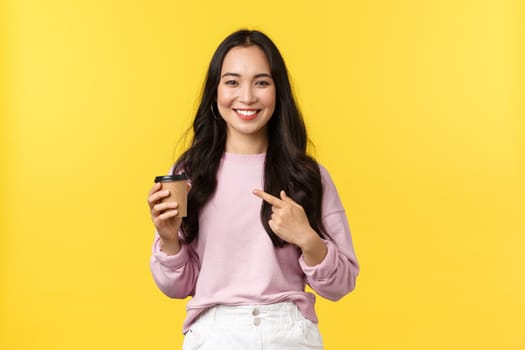 People emotions, lifestyle leisure and beauty concept. Smiling good-looking asian girl pointing at takeaway cup, recommend great cafe with delicious coffee and desserts, yellow background.