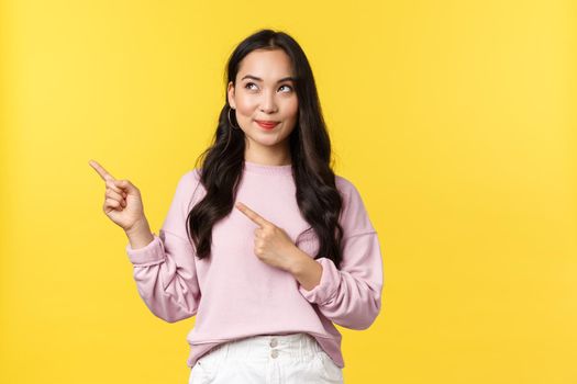 People emotions, lifestyle and fashion concept. Dreamy intrigued stylish girl found cool product in store, pointing fingers upper left corner, looking pleased and tempting, yellow background.