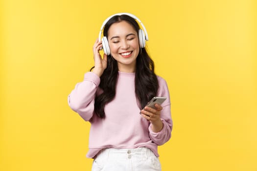 People emotions, lifestyle leisure and beauty concept. Smiling carefree modern asian girl listening music in wireless headphones. Woman enjoying favorite song in earphones, holding mobile phone.
