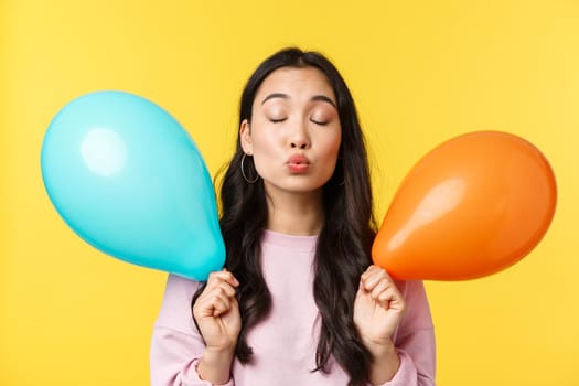 People emotions, lifestyle leisure concept. Silly and cute asian girl holding two balloons, waiting for kiss with folded lips and closed eyes, having romantic moment at party, yellow background.