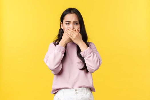 People emotions, lifestyle and fashion concept. Insulted and shocked upset asian girl cover mouth and frowning, being overwhelmed and startled over upsetting news, standing yellow background.