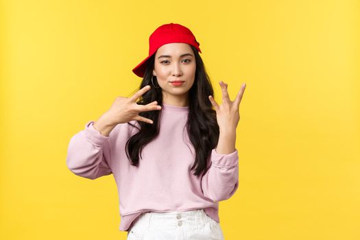 People emotions, lifestyle leisure and beauty concept. Stylish and cool, good-looking teenage korean girl in red cap, showing swag gesture standing yellow background.