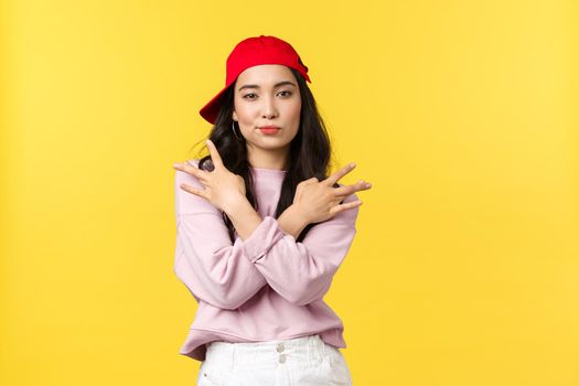 People emotions, lifestyle leisure and beauty concept. Cool and sassy stylish asian girl in red hip hop cap, showing swag gesture and smirk daring, standing confident over yellow background.