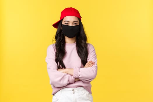 Covid-19, social-distancing lifestyle, prevent virus spread concept. Confident good-looking asian girl in red cap and face mask, cross arms chest determined, standing yellow background.