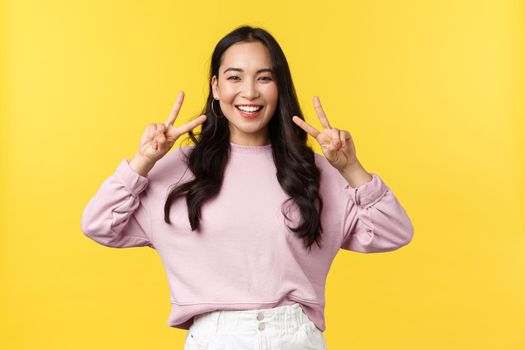 People emotions, lifestyle and fashion concept. Cheerful kawaii asian girl in stylish outfit showing peace sign and smiling upbeat with white teeth, standing over yellow background, enjoy summer.