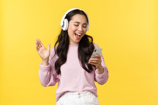 People emotions, lifestyle leisure and beauty concept. Smiling happy and carefree asian girl singing karaoke app in headphones, reading song from mobile phone, standing yellow background.