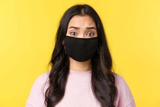 Covid-19, social-distancing lifestyle, prevent virus spread concept. Concerned and alarmed asian girl looking worried, wear face mask and frowning nervous, standing yellow background.