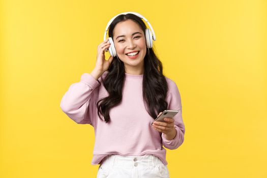 People emotions, lifestyle leisure and beauty concept. Happy smiling asian woman listening music in wireless headphones and looking pleased with nice sound, holding mobile phone.
