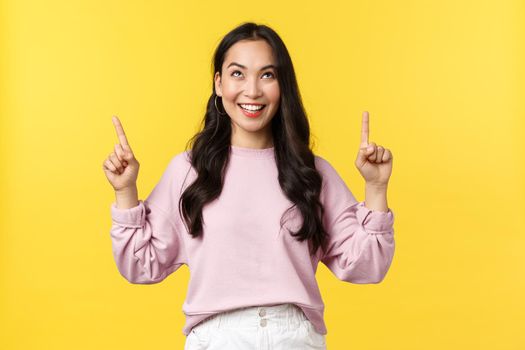 People emotions, lifestyle and fashion concept. Pleased smiling excited woman pointing fingers up and looking at top promo banner, something tasty or interesting, yellow background.