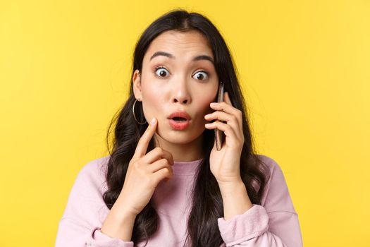 People emotions, lifestyle leisure and beauty concept. Close-up of shocked and surprised asian girl talking on phone, stare camera with popped eyes as hear big news, yellow background.