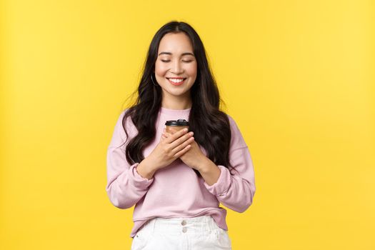 People emotions, lifestyle leisure and beauty concept. Smiling cheerful asian girl close eyes from satisfaction as enjoying favorite cup of coffee from best cafe takeaway, stand yellow background.