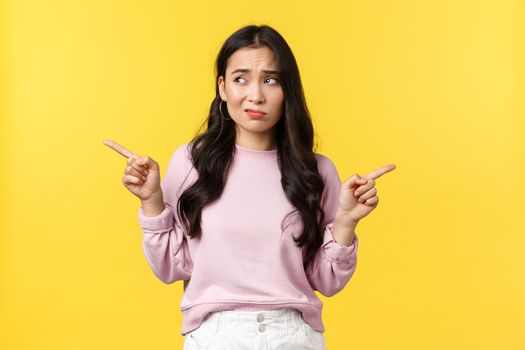 People emotions, lifestyle and fashion concept. Indecisive troubled asian woman having problem with choice, pointing fingers sideways and smirk questioned, looking left, deciding, yellow background.