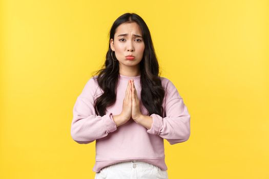 Lifestyle, emotions and advertisement concept. Pitiful cute asian girl pouting and hold hands in plead, begging for favor or help, asking for something, feeling sorry, standing yellow background.
