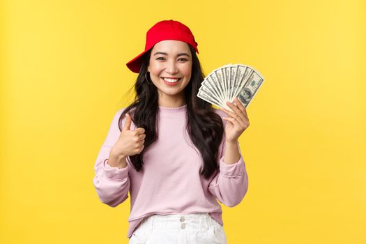 People emotions, lifestyle leisure and beauty concept. Cheerful and upbeat smiling korean girl in hip hop cap, showing thumbs-up and cash, explain how earn money on freelance, yellow background.