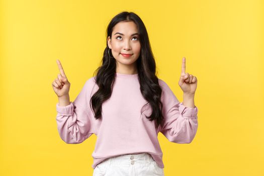 People emotions, lifestyle and fashion concept. Pleased dreamy pretty asian woman smiling and looking with temptation and desire at product upwards, pointing fingers up, yellow background.