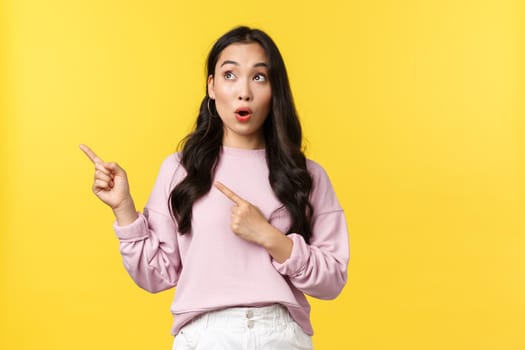 People emotions, lifestyle and fashion concept. Amazed and excited stylish asian female say wow, looking and pointing upper left corner interested in promo banner product, yellow background.